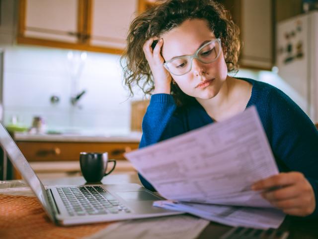 Woman seated in front of laptop holds head while reading a sheet of paper