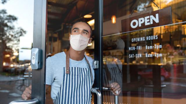 Man in apron and medical mask looks out open glass door of shop with hanging "Open" sign