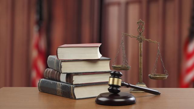 Four books stacked on a desk with a judge's gavel in the foreground and U.S. flags in the background